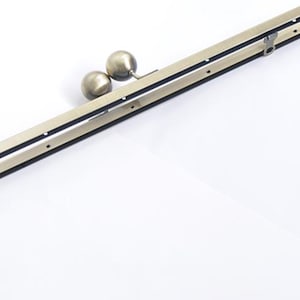 May include: A rectangular metal purse frame in a brassy gold colour. The frame has a rounded shape with a clasp featuring two small, round spheres. The frame has a black interior and small holes for sewing.