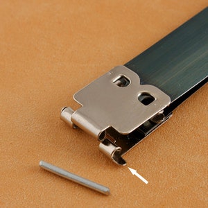 May include: A silver metal buckle with a letter "B" on it. The buckle is open and a small silver pin is lying next to it.