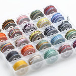 Puede incluir: Un organizador de plástico transparente lleno de 24 carretes de hilo de colores. Los colores del hilo incluyen naranja, rojo, amarillo, azul, verde y negro. Cada carrete está enrollado con una combinación de colores diferente.