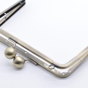 Puede incluir: Un cierre de metal dorado para un bolso o una cartera. El cierre tiene dos pomos redondos y un marco rectangular.