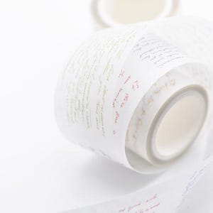 May include: Close-up of white washi tape rolls with script writing in red and green. The tape is partially unrolled, showing the inner roll and the script details. Plain white background.