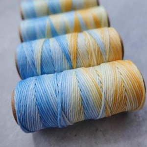 May include: Four spools of thread in a gradient of blue, yellow, and white. The thread is wound on brown cardboard spools. The colour transitions create a visually appealing effect. The spools are arranged in a row, creating a sense of depth.