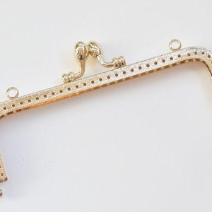 Puede incluir: Marco de bolso de metal dorado con un diseño decorativo y dos lazos en la parte superior.