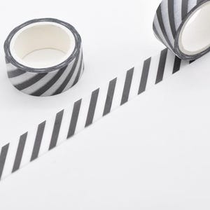 May include: Two rolls of washi tape with a black and white diagonal stripe pattern. One roll is upright, and the other is partially unrolled, with the tape extending across a white surface. The tape is a decorative craft supply.