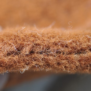May include: Close-up of a textured, brown fibre mat. The mat has a dense, interwoven structure with visible strands and a slightly uneven surface. The fibres are a warm, earthy brown colour.