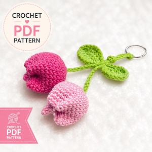 May include: A crochet keychain featuring two tulip-shaped charms in dark pink and light pink yarn, attached to green stems with two green leaves and a silver metal ring. The image includes text that reads CROCHET PDF PATTERN in two locations.