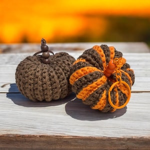 May include: Two crocheted pumpkin decorations. One is solid brown, and the other is brown with orange stripes. Both have brown stems and orange keychains. The pumpkins are sitting on a light-colored wooden surface.