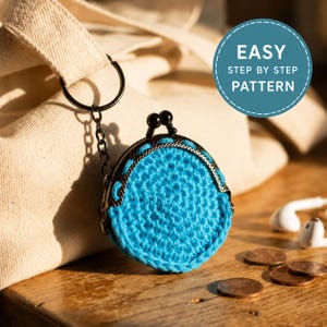May include: A round, bright blue crocheted coin purse with a black metal kiss-lock frame and a matching black keychain attachment. A circular teal graphic overlay in the upper right corner contains the white text EASY STEP BY STEP PATTERN.