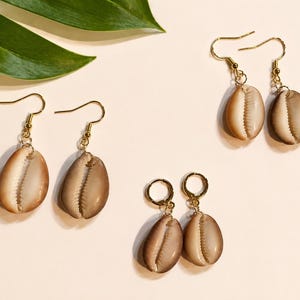 May include: Four pairs of seashell earrings with gold-colored hooks and hoops are displayed on a light background. The earrings feature natural-colored, oval-shaped shells. A green leaf is in the upper left corner.