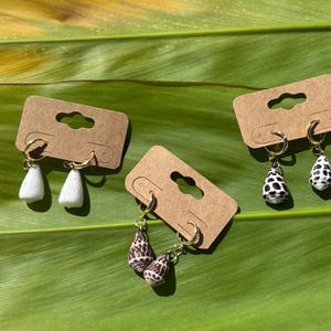 May include: Three pairs of hoop earrings attached to brown cardboard display cards. One pair features solid white cone-shaped shells, another has dark brown patterned shells, and the third pair displays white shells with black spots.
