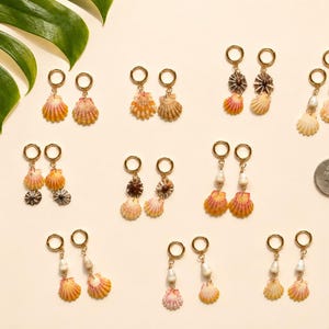 May include: A collection of seashell earrings with gold-colored hoops and various designs. Some earrings feature multiple shells, while others include pearls. The shells display shades of orange, pink, and cream, with a quarter coin for size reference.