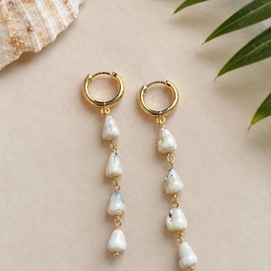 May include: A pair of gold hoop earrings featuring dangling chains adorned with four small, white, irregularly shaped stone beads each. The beads have subtle grey and black speckles and are connected by thin gold metal links.