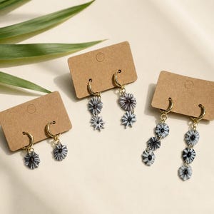 Könnte beinhalten: Drei Paar Ohrringe auf braunen Ohrringkarten. Die Ohrringe haben goldene Reifen und ein hängendes Design mit kleinen, runden, schwarz-weiß gemusterten Anhängern. Die Ohrringe werden auf einer hellen Oberfläche mit grünen Blättern präsentiert.
