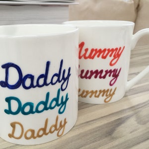 May include: Two white ceramic mugs with handles. The mug on the left has the word "Daddy" written three times in blue, teal, and gold. The mug on the right has the word "Mummy" written three times in red, purple, and gold.