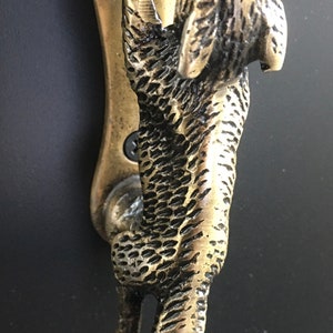 Pode incluir: Um batedor de porta de latão em forma de raposa segurando uma bola nas patas. A raposa é detalhada com pêlo texturizado e tem uma aparência realista.