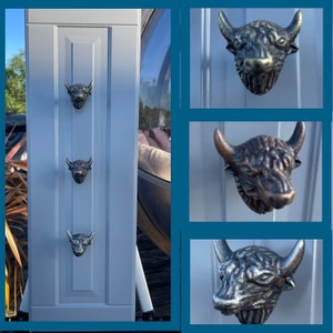 May include: A pale grey cabinet door featuring three bronze-toned bull head knobs. The knobs are arranged vertically. The door has vertical panel detailing.