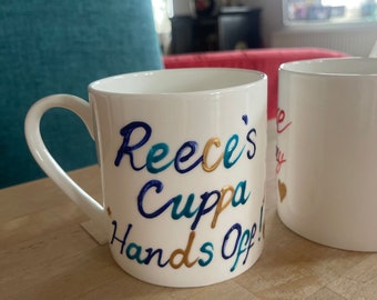 May include: White ceramic mug with the words "Reece's Cuppa Hands Off!" written in colorful, raised lettering. The mug has a blue, gold, and teal color scheme.