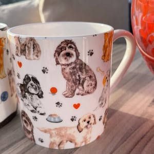 以下が含まれることがあります： 茶色、黒、白の色合いで様々な犬種が描かれた、繰り返し模様の白いセラミック製マグカップ。表面には小さな赤いハート、オレンジ色のボール、犬の足跡、骨、フードボウルが散りばめられたデザインが施されています。