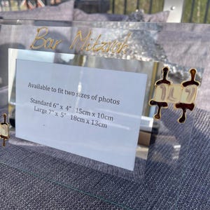 May include: A clear acrylic picture frame featuring gold-colored text reading Bar Mitzvah at the top and two decorative Torah scroll icons on the sides. A white insert displays text regarding photo sizes: Standard 6" x 4" and Large 7" x 5".