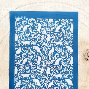 May include: A blue stencil with a white design of cats and floral patterns. The stencil has the text "moiko 20.22" in the top left corner and "moiko" in the bottom right corner.