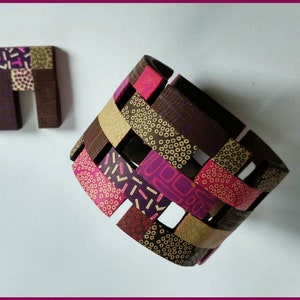 K&ouml;nnte beinhalten: Ein gewebtes Armband aus Polymer Clay mit einem geometrischen Muster in Rosat&ouml;nen, Braun und Gold. Das Armband besteht aus einzelnen Tonst&uuml;cken, die miteinander verwoben sind, um ein einzigartiges und komplexes Design zu schaffen. Das Armband wird au&szlig;erdem von einem kleinen, quadratischen Tonst&uuml;ck mit einem &auml;hnlichen geometrischen Muster begleitet.
