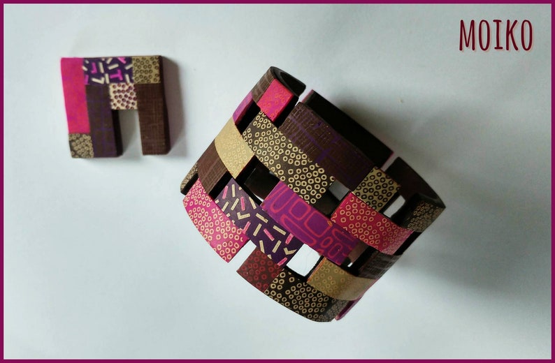 K&ouml;nnte beinhalten: Ein gewebtes Armband aus Polymer Clay mit einem geometrischen Muster in Rosat&ouml;nen, Braun und Gold. Das Armband besteht aus einzelnen Tonst&uuml;cken, die miteinander verwoben sind, um ein einzigartiges und komplexes Design zu schaffen. Das Armband wird au&szlig;erdem von einem kleinen, quadratischen Tonst&uuml;ck mit einem &auml;hnlichen geometrischen Muster begleitet.