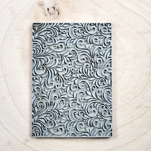 Op de afbeelding: Een grijze rubberen stempel met een gedetailleerd bloemenpatroon. Het patroon heeft ingewikkelde swirls en bladeren.