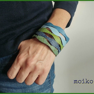 K&ouml;nnte beinhalten: Eine Person tr&auml;gt ein blaues Hemd und Jeans mit einem Stapel von f&uuml;nf bunten, gemusterten Armb&auml;ndern am Handgelenk. Die Armb&auml;nder sind gr&uuml;n, blau und wei&szlig;.