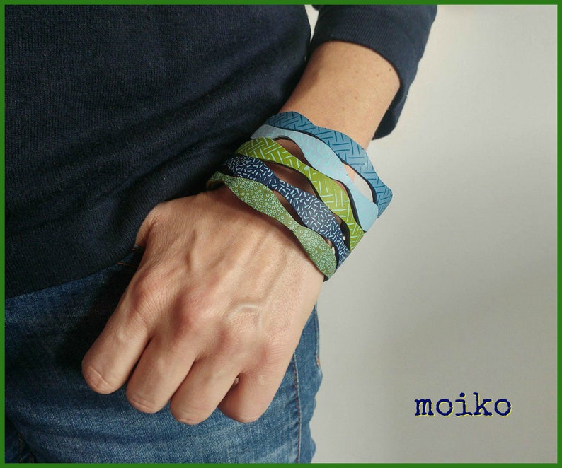 K&ouml;nnte beinhalten: Eine Person tr&auml;gt ein blaues Hemd und Jeans mit einem Stapel von f&uuml;nf bunten, gemusterten Armb&auml;ndern am Handgelenk. Die Armb&auml;nder sind gr&uuml;n, blau und wei&szlig;.