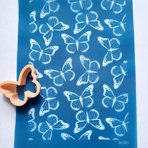 Op de afbeelding: Een blauwe siliconen bakmat met een wit vlinderpatroon. De mat heeft de tekst "moiko 10.05" in de linkerbovenhoek en "moiko" in de rechterbenedenhoek. Een plastic vlinderkoekjesvormpje bevindt zich in de linkerbenedenhoek.