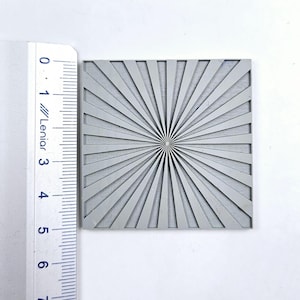 Puede incluir: Una plantilla cuadrada gris con un diseño de rayos de sol radiantes. La plantilla mide aproximadamente 5 cm de ancho.