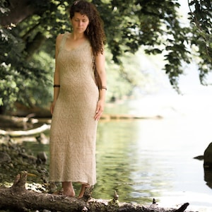 May include: A woman wearing a long, beige, knitted dress stands on a log by a lake. The dress has a chevron pattern and the woman is barefoot.