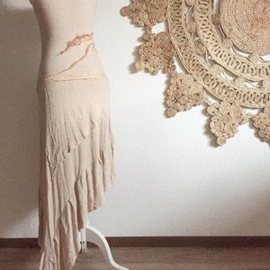 May include: A long, flowing, light brown dress with a ruffled hem and a brown design on the bodice. The dress is displayed on a white mannequin against a white wall.