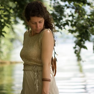 May include: A woman wearing a light brown crocheted top and skirt with a brown bracelet on her wrist. She is standing in front of a body of water with trees in the background.