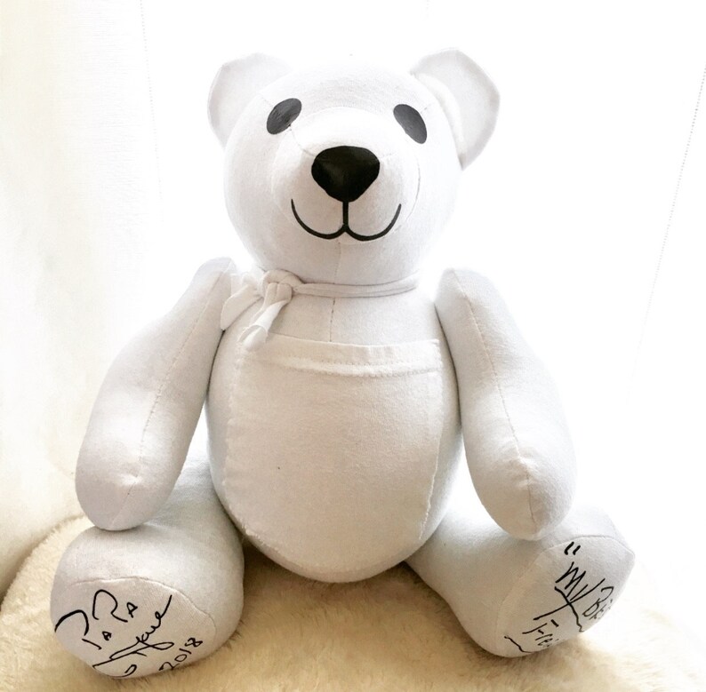 May include: A white fabric teddy bear with black embroidered eyes and a black nose. The bear features a small fabric tie around its neck and a pocket on its chest. Handwritten black text is visible on both feet, including the year 2018.