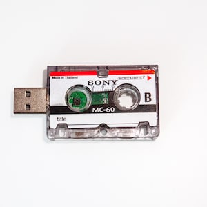 Puede incluir: Una cinta de cassette de plástico transparente con una unidad USB plateada adjunta. La cinta de cassette está etiquetada como "SONY" y "MC-60" y tiene una etiqueta roja y blanca con el texto "MICROCASSETTE".