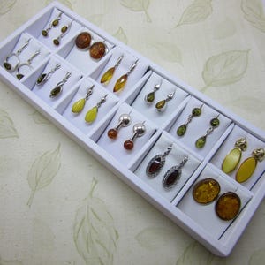 May include: A white display tray with 10 pairs of amber earrings. The earrings are in various shapes and sizes, including teardrop, oval, and round. The earrings are all silver.