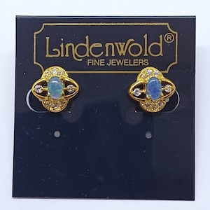 May include: A pair of gold earrings featuring opal gemstones and small clear cubic zirconia stones. The earrings are displayed on a black card with the text "Lindenwold Fine Jewelers" printed on it.