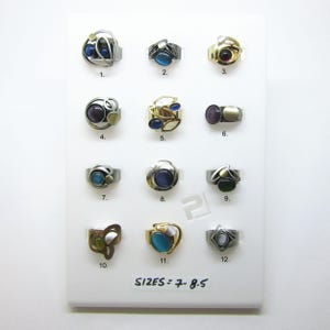 May include: A collection of twelve rings with various designs and gemstones, displayed on a white surface. The rings feature silver, gold, and mixed metal bands, with stones in shades of blue, purple, green, and white. Sizes range from 7 to 8.5.