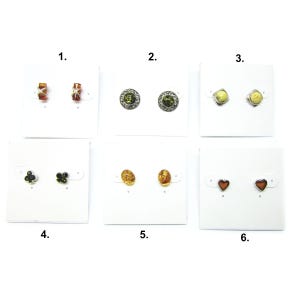 May include: Six pairs of stud earrings are presented on white cards. The earrings showcase diverse designs, including amber, green stones, and heart shapes. Some have silver settings, while others feature distinctive patterns.