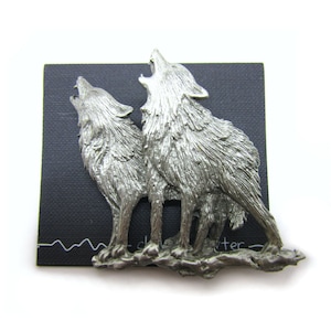 May include: A silver pewter pin featuring two wolves howling at the moon. The wolves are standing on a rocky base.