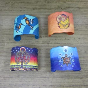 May include: Four colorful fabric bracelets with different designs. The bracelets are blue, orange, and purple with images of birds, a dream catcher, a tree, and a turtle.