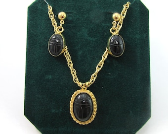 May include: A gold-toned necklace and earring set. The necklace features a pendant with a black oval stone, and the earrings have matching black oval stones. The set is displayed on a dark green velvet background.