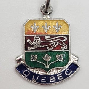 May include: A silver-toned Quebec charm with a colorful enamel design. The charm features a shield shape with a yellow top, a red and black center with a lion, and a green and blue bottom with the word "QUEBEC".