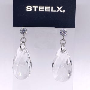 Può includere: Un paio di orecchini pendenti in cristallo trasparente con una scintillante borchia rotonda e un pendente in cristallo a forma di goccia. Gli orecchini sono sospesi da un anello in metallo color argento. Il marchio "STEELX" è stampato in alto.