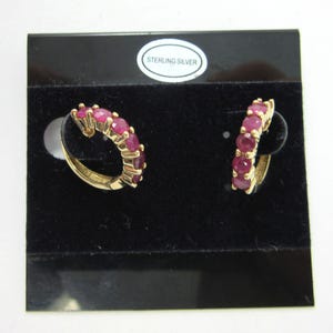 May include: A pair of gold-coloured hoop earrings, each featuring a line of bright red gemstones. The earrings are presented on a black velvet surface, with a "STERLING SILVER" sticker in the background.