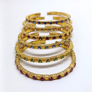 Puede incluir: Un conjunto de cinco pulseras doradas con brillantes de colores. Las pulseras están apiladas una encima de la otra, con la pulsera inferior con brillantes rosas, la siguiente con verdes, luego azules, blancos y finalmente azules.