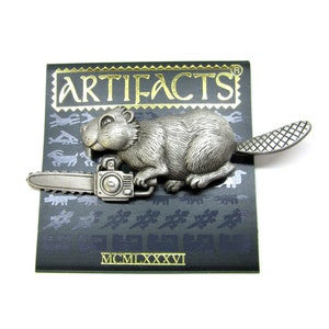 May include: A silver pewter pin of a beaver holding a chainsaw. The pin is on a black card with gold lettering that reads "ARTIFACTS" and "MCMILXXXVI".