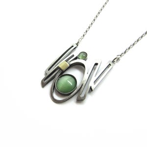 Puede incluir: Un collar de metal plateado con un colgante geométrico abstracto de barras de marco abierto. El colgante está acentuado con un cabujón verde redondo grande, una cuenta verde redonda más pequeña y una pequeña pieza rectangular de color amarillo pálido.