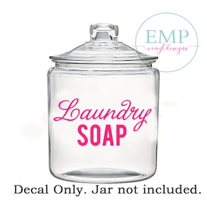 Puede incluir: Un tarro de cristal transparente con tapa, con las palabras "Laundry SOAP" en escritura rosa brillante. La calcoman&iacute;a est&aacute; dise&ntilde;ada para la organizaci&oacute;n del lavadero. La imagen tambi&eacute;n incluye un logotipo en un c&iacute;rculo que dice "EMP vinyl designs".